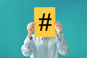 A person in a light blue shirt holds up a bright yellow sign with a large black hashtag symbol, questioning, Should Hashtags Be Capitalized? against a vibrant turquoise background. The persons face is obscured by the sign.