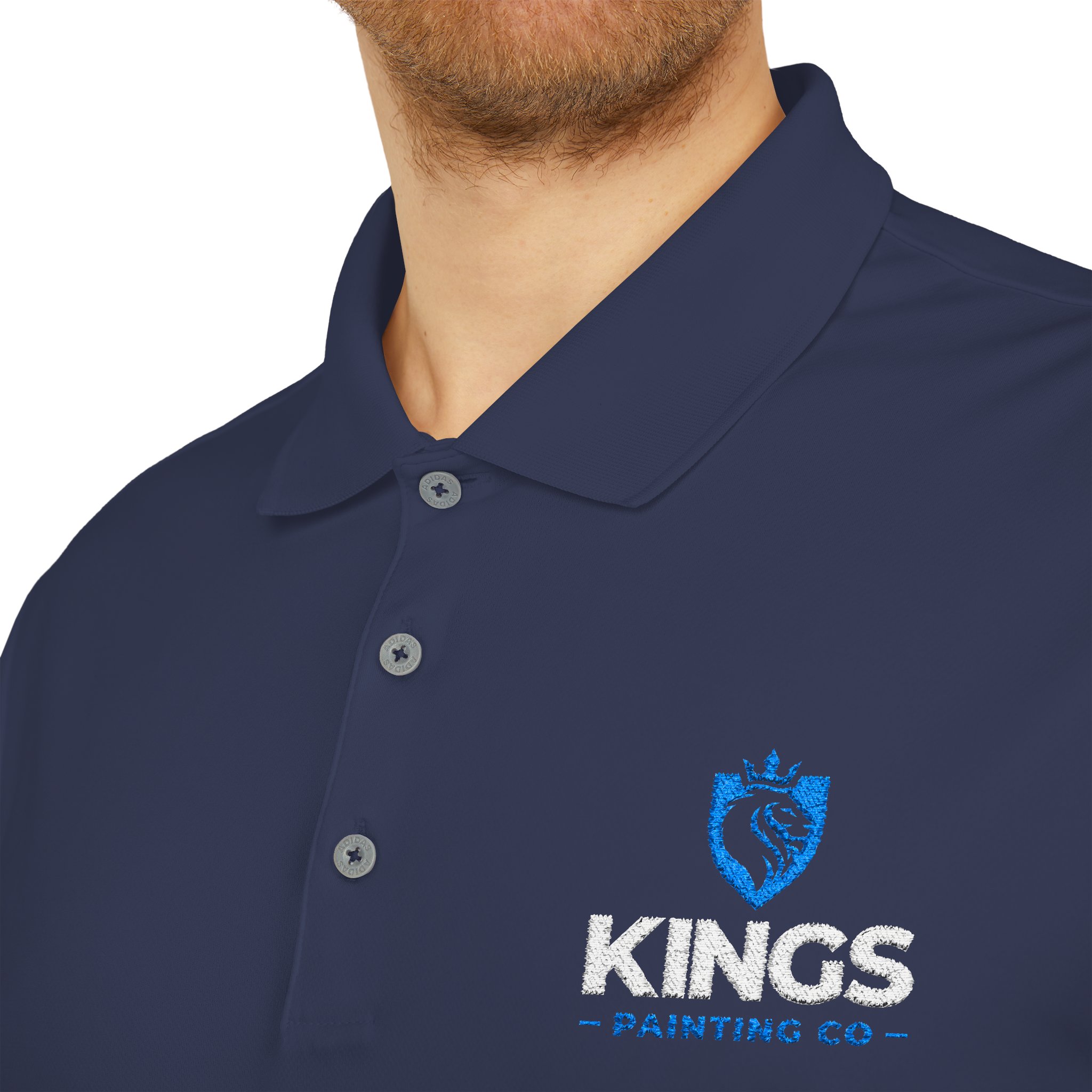 Close-up of a person wearing the Kings Painting Co. Performance Polo by Under Armour®, featuring the KINGS PAINTING CO logo with a crowned lion inside a shield embroidered in white and blue on the chest.