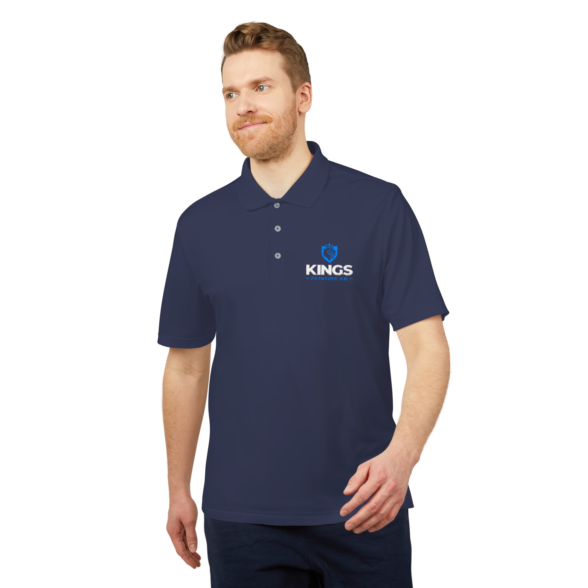 A man with light hair and a beard is wearing the Kings Painting Co. Performance Polo by Under Armour®, which features “KINGS” and a blue lion logo on the chest. He is standing against a white background, looking slightly to the side.