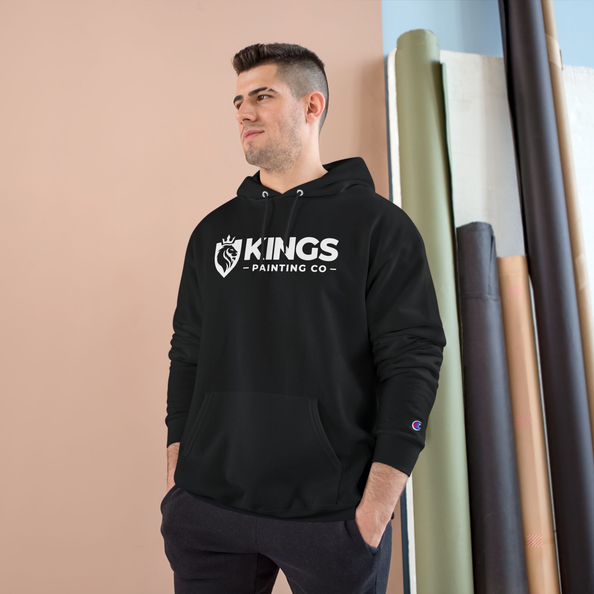 A man wearing the Kings Painting Co. Logo Hoodie by Champion® stands indoors near tall rolls of fabric or paper, looking off to the side with a slight smile.